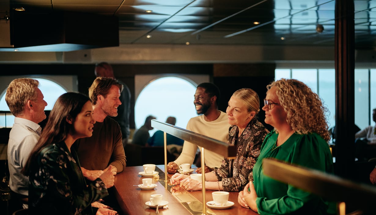 Gruppe af mennesker der sidder ved en disk og drikker kaffe, med store vinduer i baggrunden