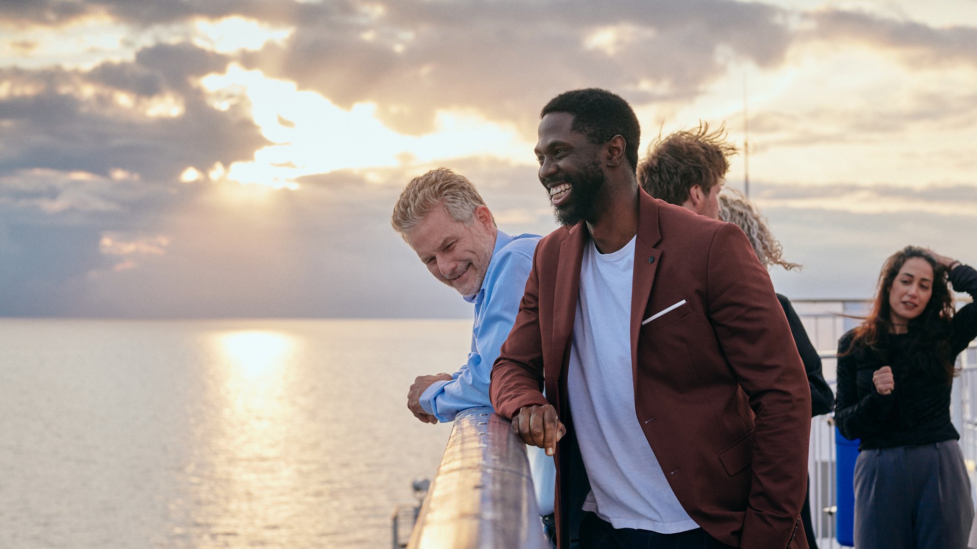 Fire personer, der står på en terrasse med havet og solnedgang i baggrunden.