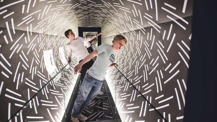 To personer går gennem en oplyst tunnel med hvide linjemønstre.