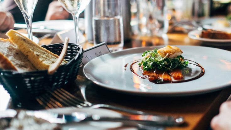 Spisebord med gourmetret, vinglas og brødstænger i en formel restaurantopsætning.