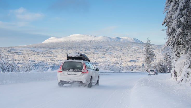 Hvid bil der kører på en snedækket vej med bjerge i baggrunden.
