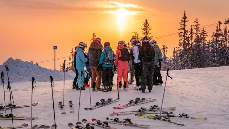 Gruppe mennesker på en snedækket bakke med skiudstyr ved solnedgang.