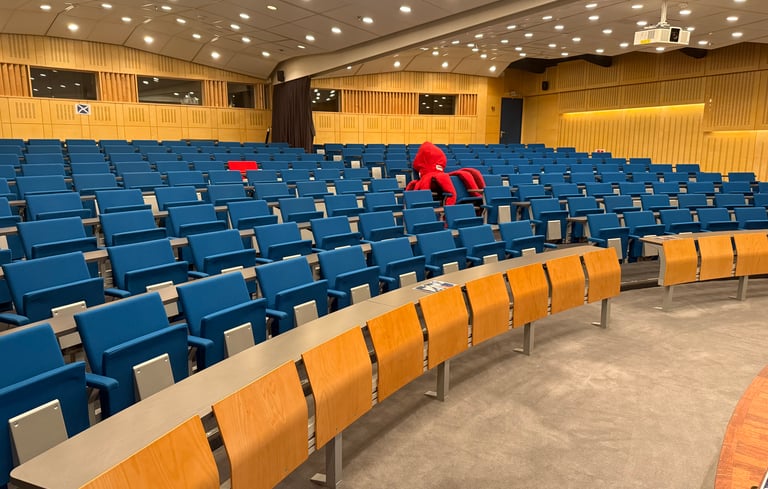 Tomt auditorium med blå sæder og træborde, én person i rødt sidder alene