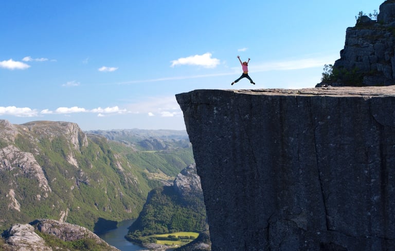 Person hopper på klippekant med udsigt over landskab med bjerge og flod.