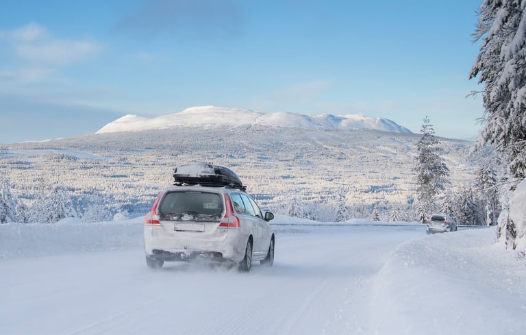 Hvid bil der kører på en snedækket vej med bjerge i baggrunden.