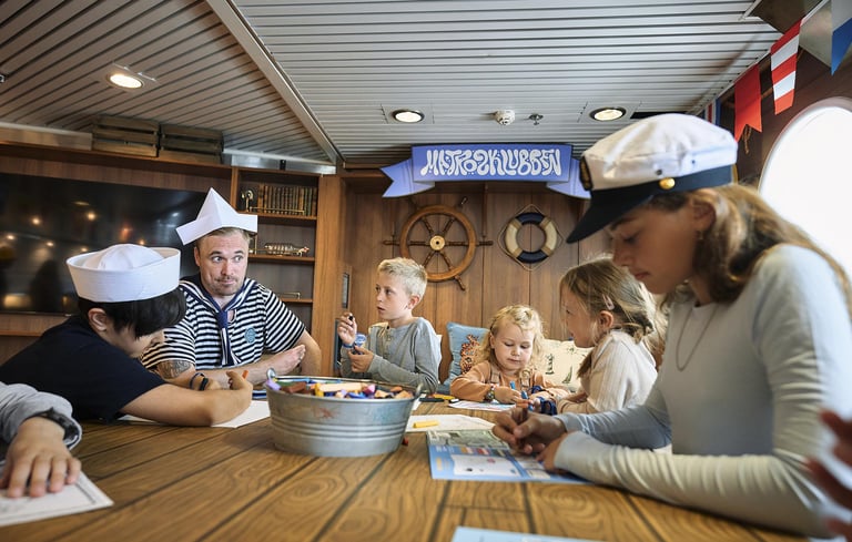 Gruppe af mennesker, herunder børn og voksne, der deltager i en aktivitet med papir og markører i Matrosklubben.